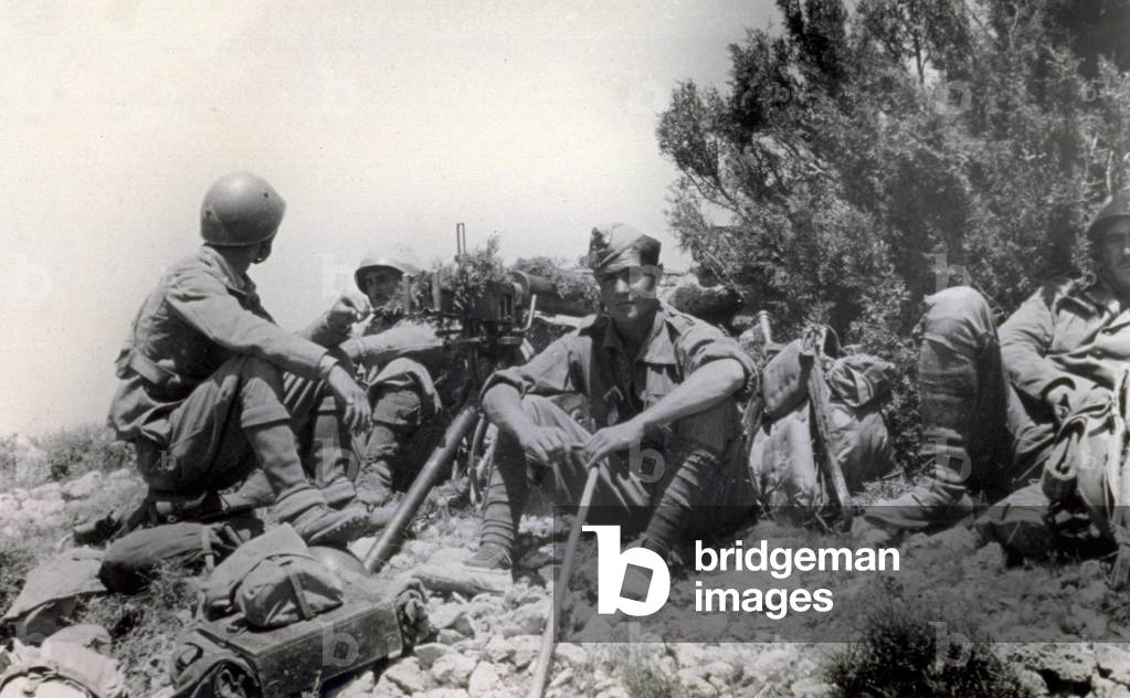 A few Italian soldiers of the sixth squadron of the Machine Gun Battalion resting in the emplacement on July 13, 1938, during the action known as 'Del Levante'. The men are sitting on the ground with their equipment backpacks and a machine gun set up next to them (b/w photo)