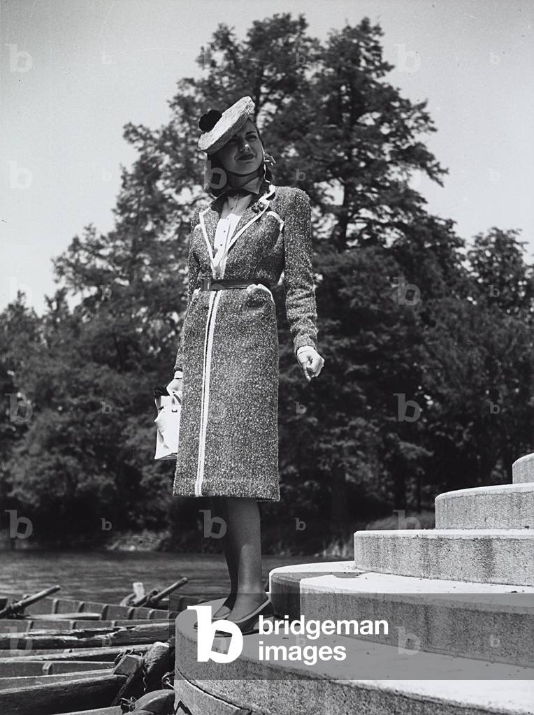 A model posing on the shore of a lake wearing a wool coat and a hat with a pom-pom. (b/w photo)