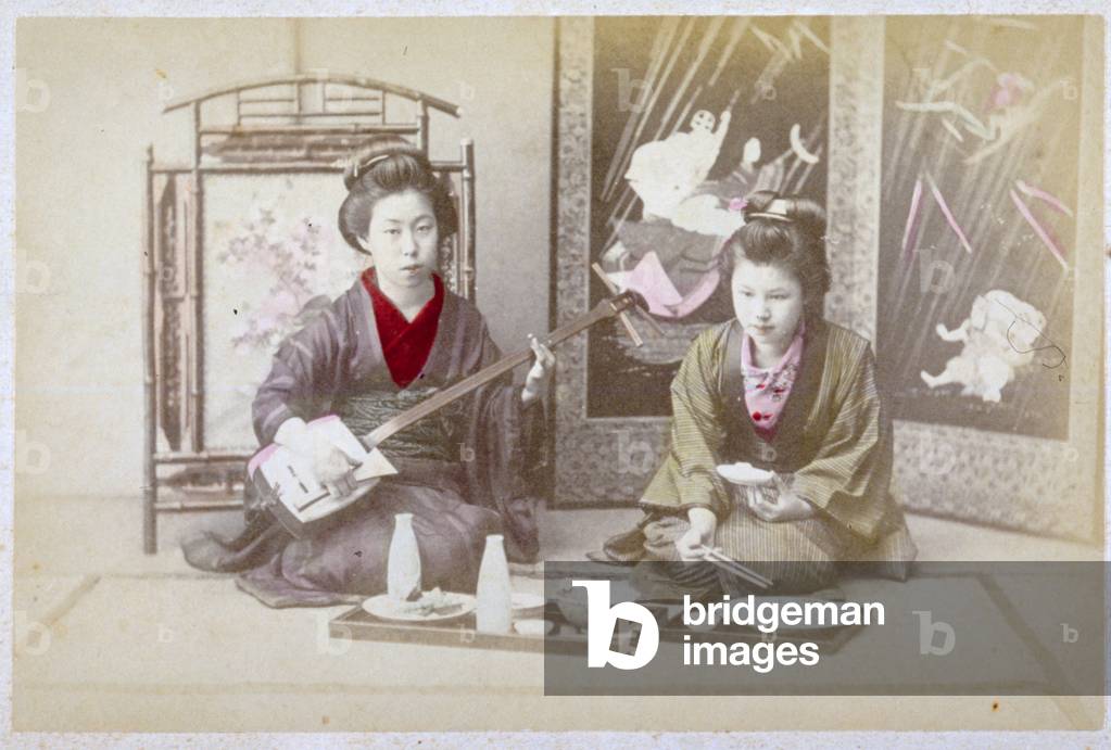 Full-length portrait of two young Japanese women sitting on the ground. They are wearing traditional clothes. The young woman to the left plays the Syamisen, the other is eating, helping herself to the food from two trays laid out on the ground, in front of them