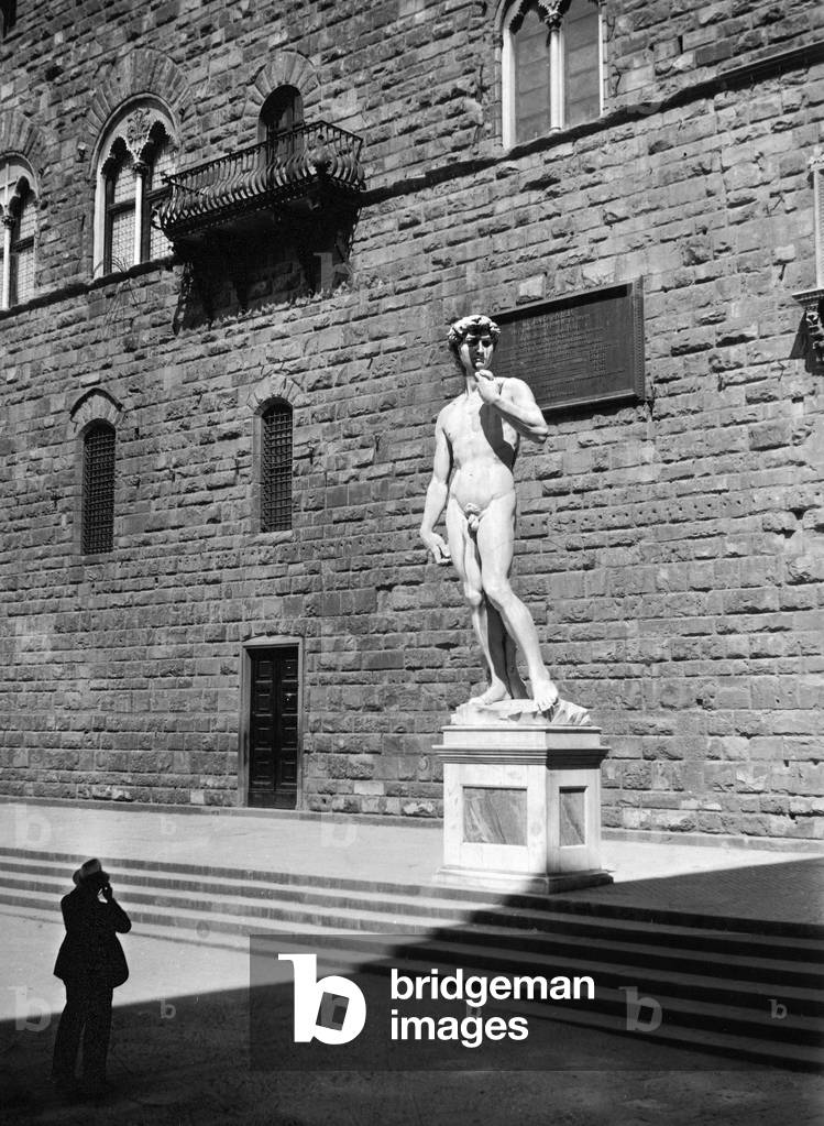 Foreshortened image of Signoria Place in Florence, the David by Michelangelo and part of Palazzo Vecchio can be seen. In the foreground a man taking a photograph.