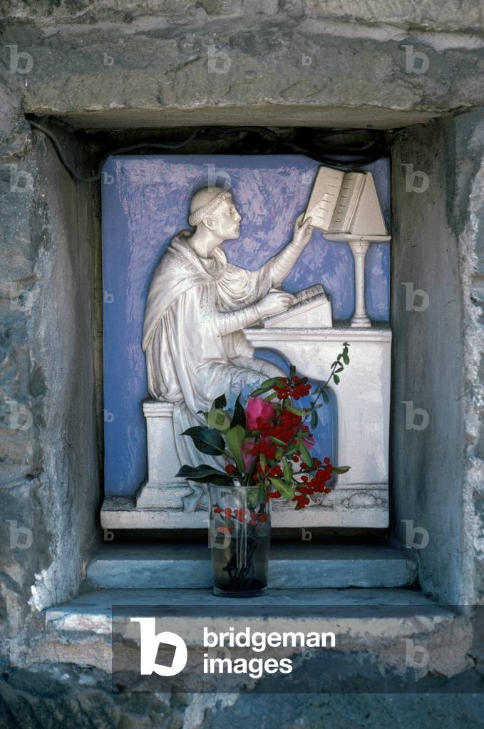 Bas-relief in glazed terra cotta of a monk seated at a desk copying a text. The work is on the outer wall of a building in Consuma, in the Tosco-Romagnolo Apennines, Italy (photo)