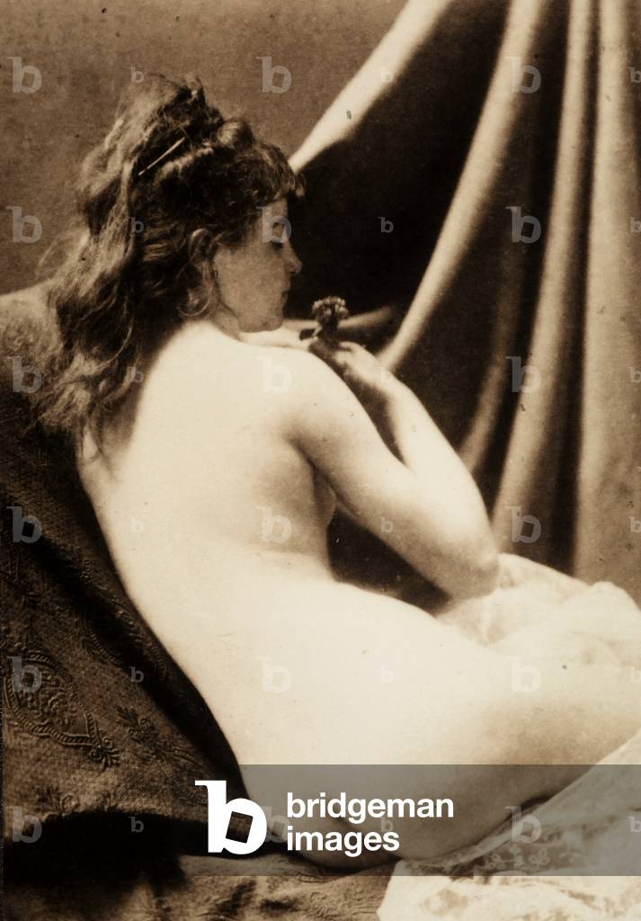 Nude portrait of a young woman seen from the rear, seated, as she smells a flower.