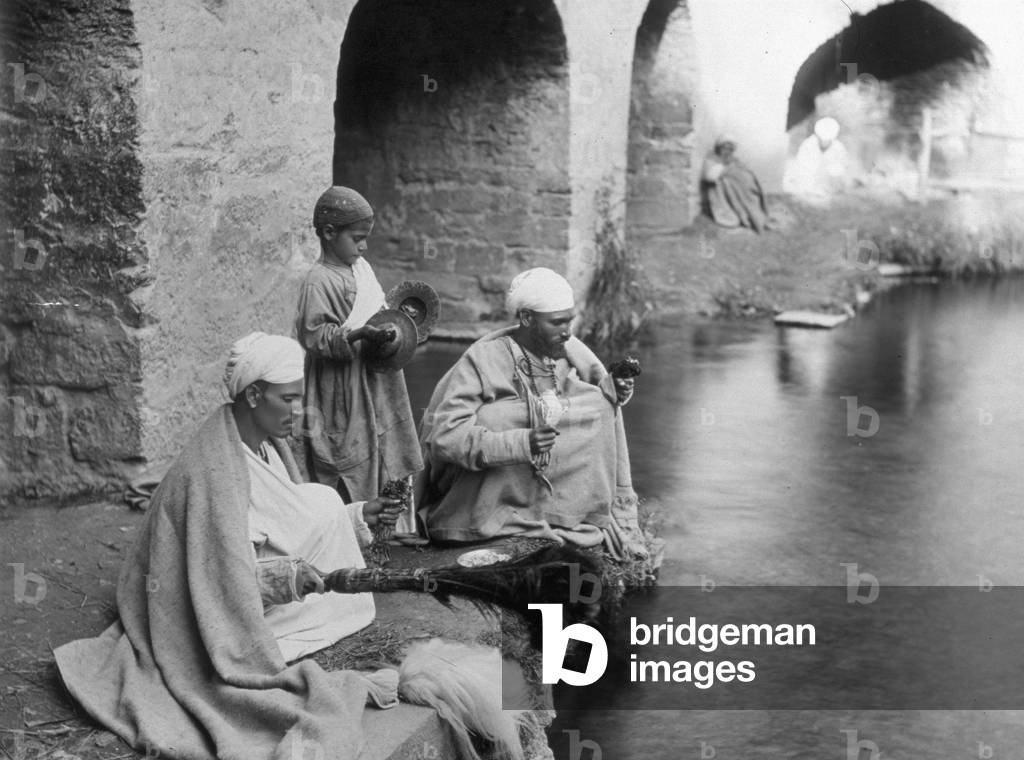 Two Indians from Kashmir shown with a child on the banks of a river. They are wearing traditional attire and are performing a rite facing the great river, 1900 (print on double-weight paper)