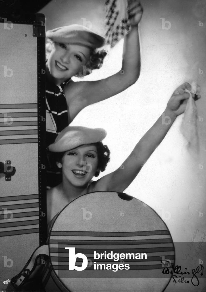 Two soubrettes waving and smiling behind a trunk and a hatbox