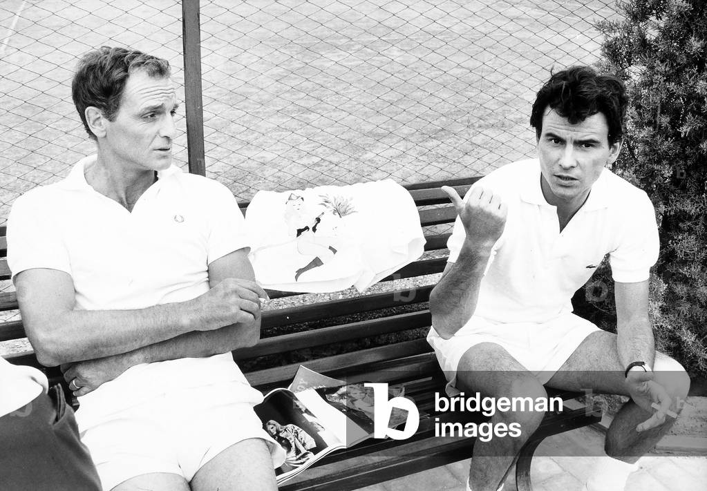 Portrait of the actors Philippe Leroy and Horst Bucholz wearing tennis outfits. They are smoking on a bench, between them are some magazines.