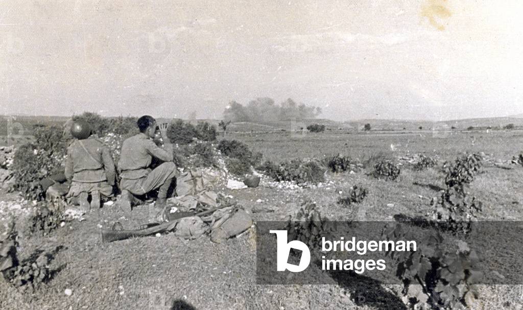 Two Italian soldiers, kneeling on the ground, observing the bombings carried out by the Italian Airforce during the Spanish civil war, on July 14, 1938, in the course of the action known as 'Del Levante'. In the distance columns of smoke caused by the explosions. The landscape is almost level, with bushes. In the foreground the backpacks with military equipment and a rifle are set on the ground (b/w photo)