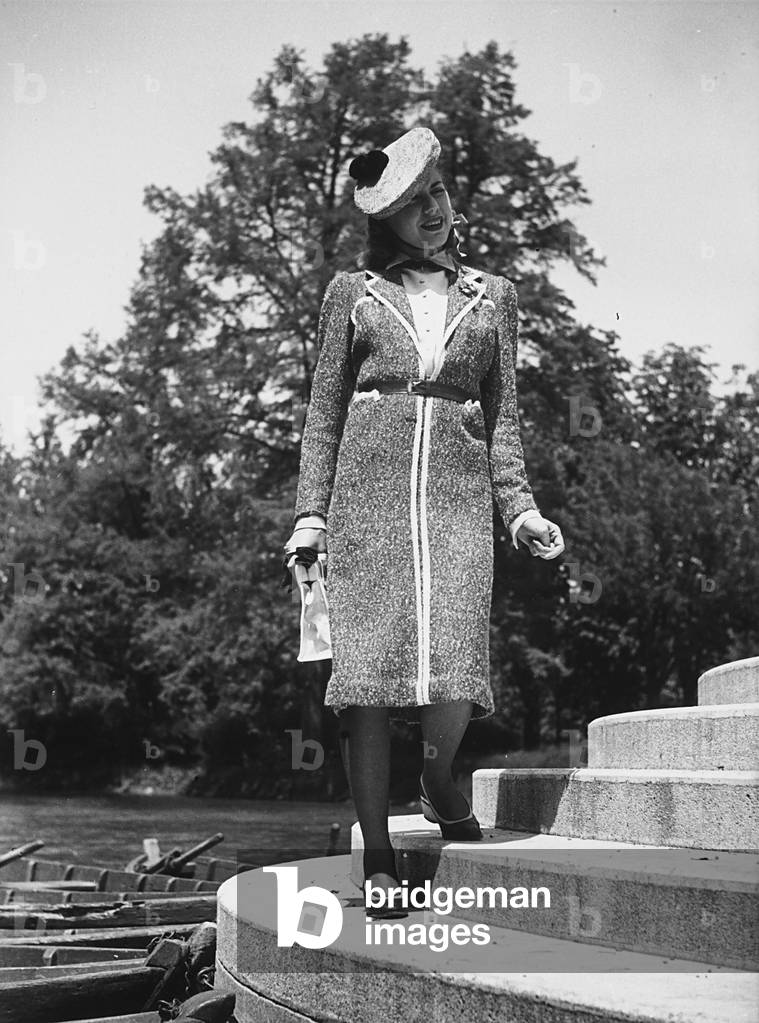 Model posing and smiling on the shore of a lake wearing a wool coat and a hat with a pom-pom. (b/w photo)