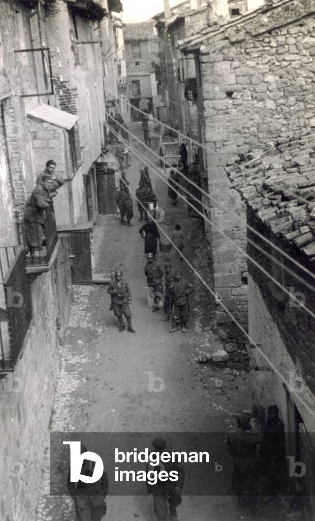 View of a street in Horta de S. Juan taken from the top of a building. Soldiers in uniform and a few civilians can be seen walking along the street (b/w photo)