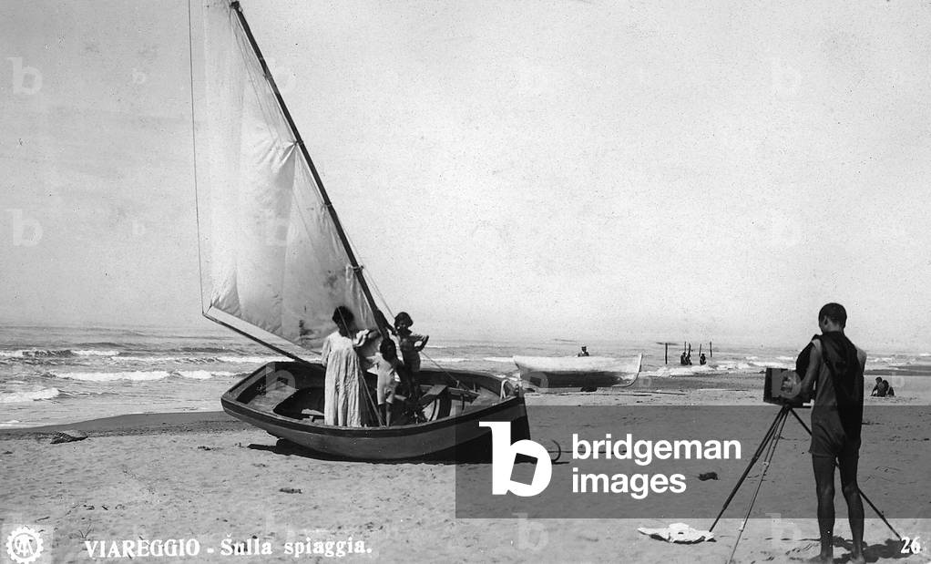 A photographer, with his back to the viewer, focuses his camera on a woman and two children, standing in a small boat on the shore of a beach in Viareggio.