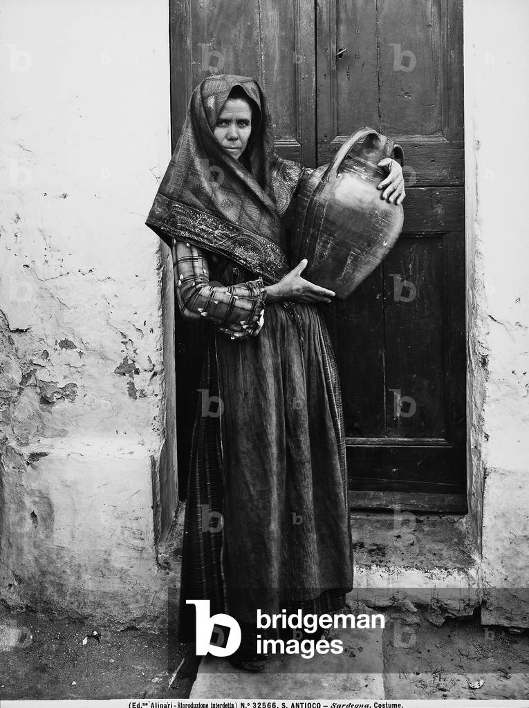 Woman in traditional dress, with an amphora in her arms, photographed in the door of a house on the island of Sant'Antioco.