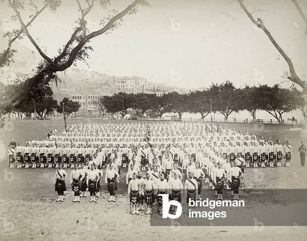 The image shows a troop of the Royal British Army, lined up in more rows on a grass of a park, near to Hong Kong's harbour. The soldiers wear the Scottish uniform.
