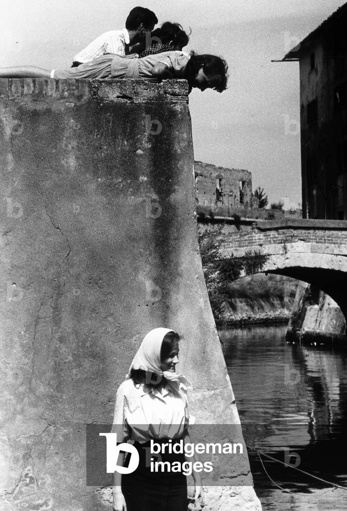 Carla Gravina smiling with a kerchief tied around her head along the shore of a river. Part of a bridge, and a nearby wall, two children and an adult are visible.