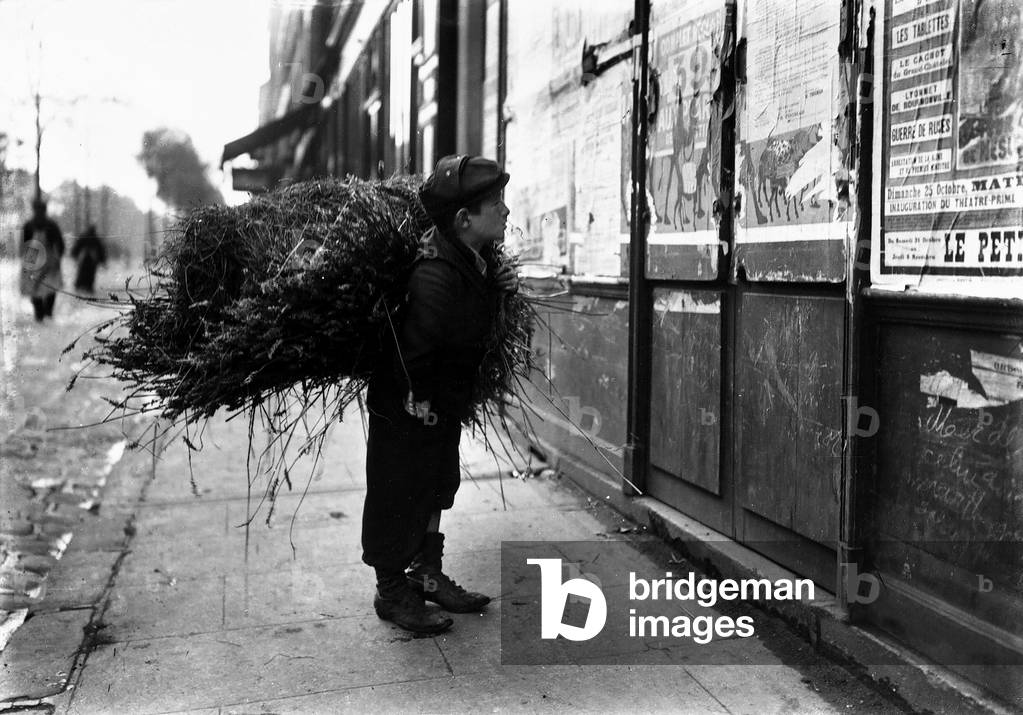 Child with a bundle of sticks on his shoulder looking at a poster affixed to a door in Paris.