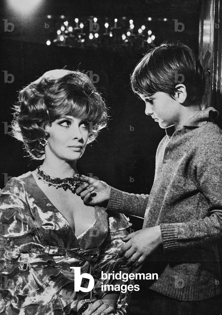 The actress Gina Lollobrigida sitting next to her son Mirko junior. He is touching and admiring her necklace.