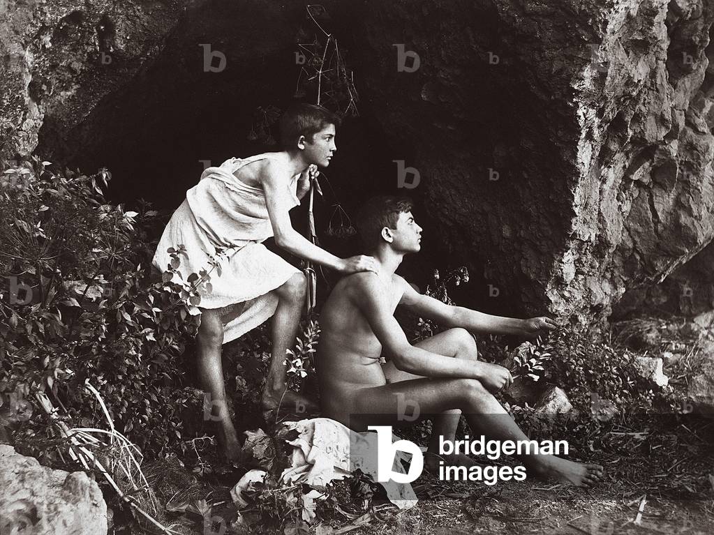 Artistica photograph invoking the Arcadian atmosphere of the ancient Greece. An adolescent nude in profile, is sitting in front of a grotto. Behind the youth is a boy dressed as a shepherd