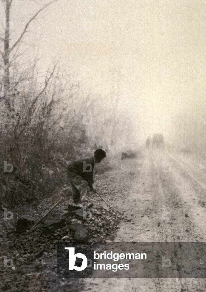 A country road wrapped in morning mists. In the foreground a man is splitting stones