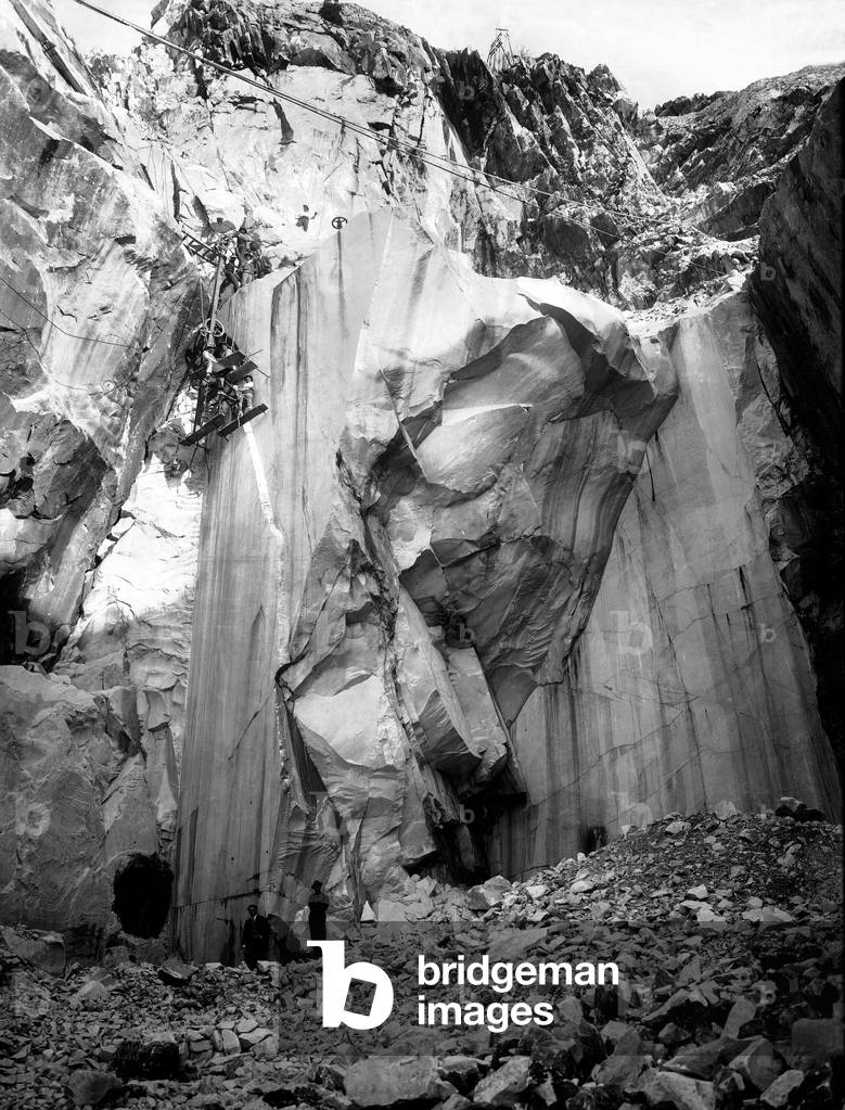 Excavators at work along a steep wall of a marble quarry in the Apuan Alps, Tuscany. (b/w photo)