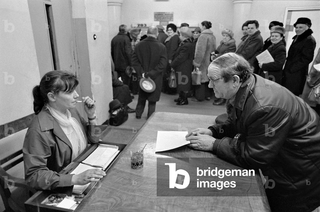 Warsaw, Poland, 1979. Queue in public office. Female clerk smokes cigarettes while talking with inquirer. 