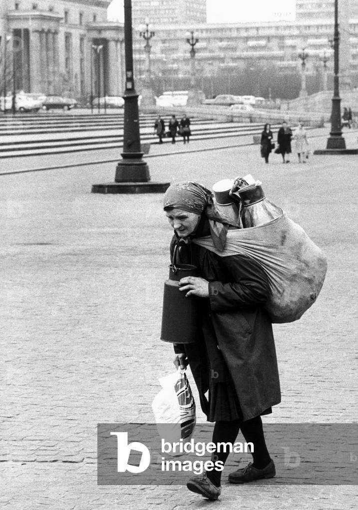 Warsaw, Poland, 1973. Old woman carrying a sack with milk cans on her back near the Palace of Culture and Science. 