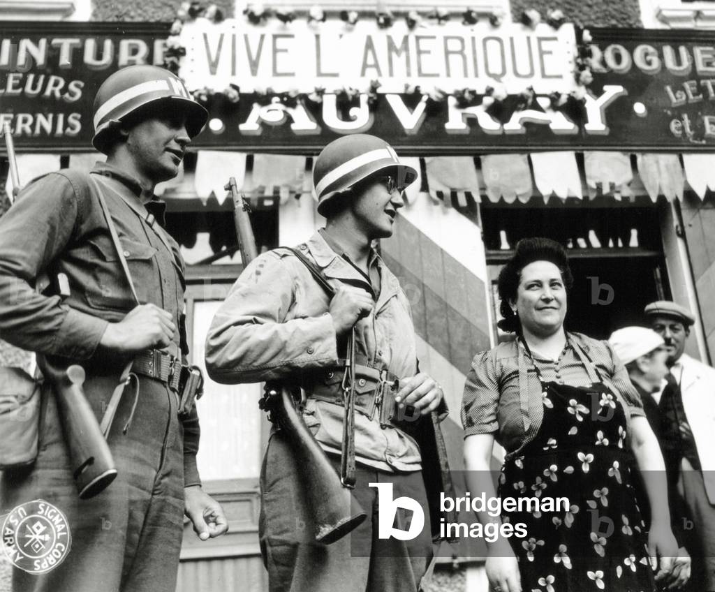 Two U.S. Military Police stand with a woman under the name of the paint-drugstore A. Auvray that displays a sign 'Vive L'Amerique' (Long live America), Rue Balleroy, Molay-Littry, Normandy, France, June 1944 (b/w photo)