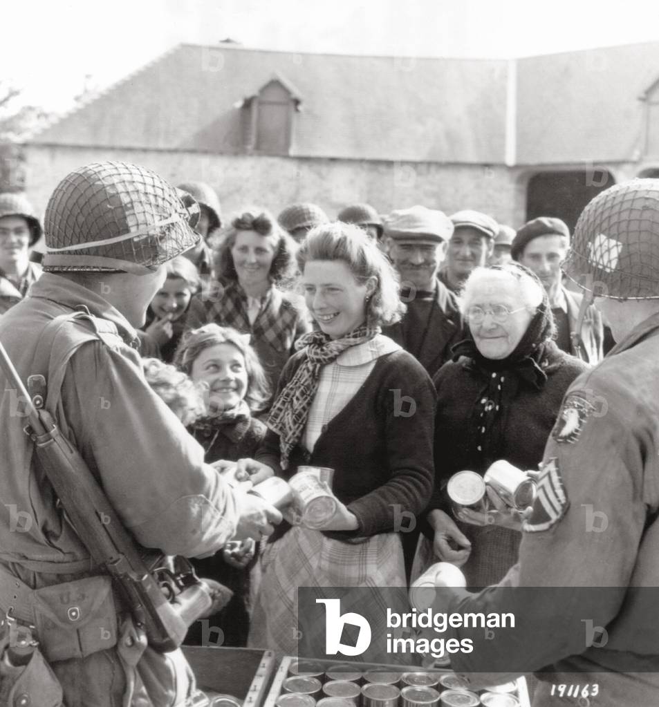 American paratroopers of the 801st Ordnance Company of the 101st  Airborne Division are distributing food (cans) to civilians in the courtyard of the farm Lecaudey Hiesville, Hiesville, Normandy, France, June 1944 (b/w photo)