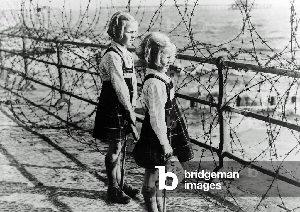 Two girls on the south coast of England look out toward the beach through a barbed wire fence constructed as part of Britain's coastal defenses, 1940 (b/w photo)