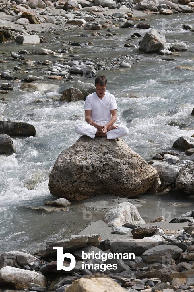 Man meditating in the lotus position, sitting on a rock by the river, Les Contamines, France