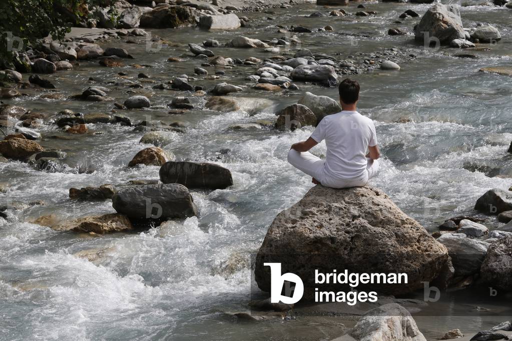 Man meditating in the lotus position, sitting on a rock by the river, Les Contamines, France