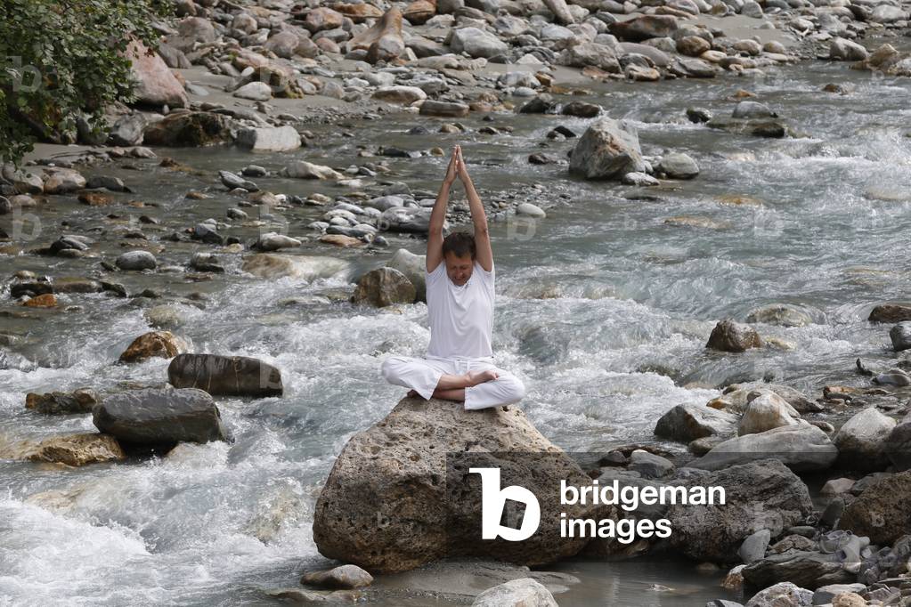 Man meditating in the lotus position, sitting on a rock by the river, Les Contamines, France