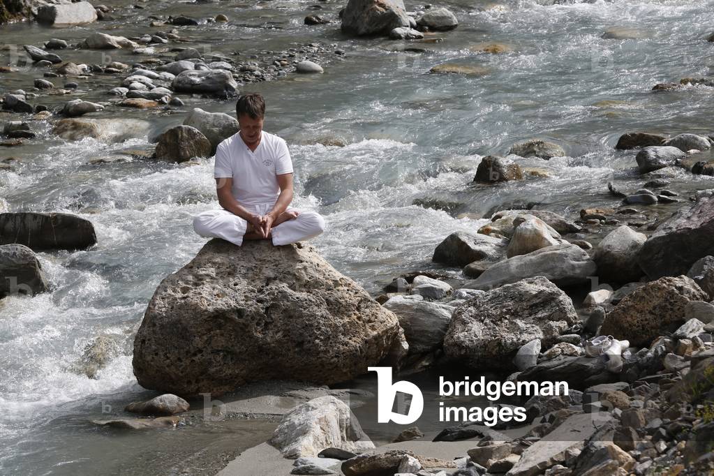 Man meditating in the lotus position, sitting on a rock by the river, Les Contamines, France