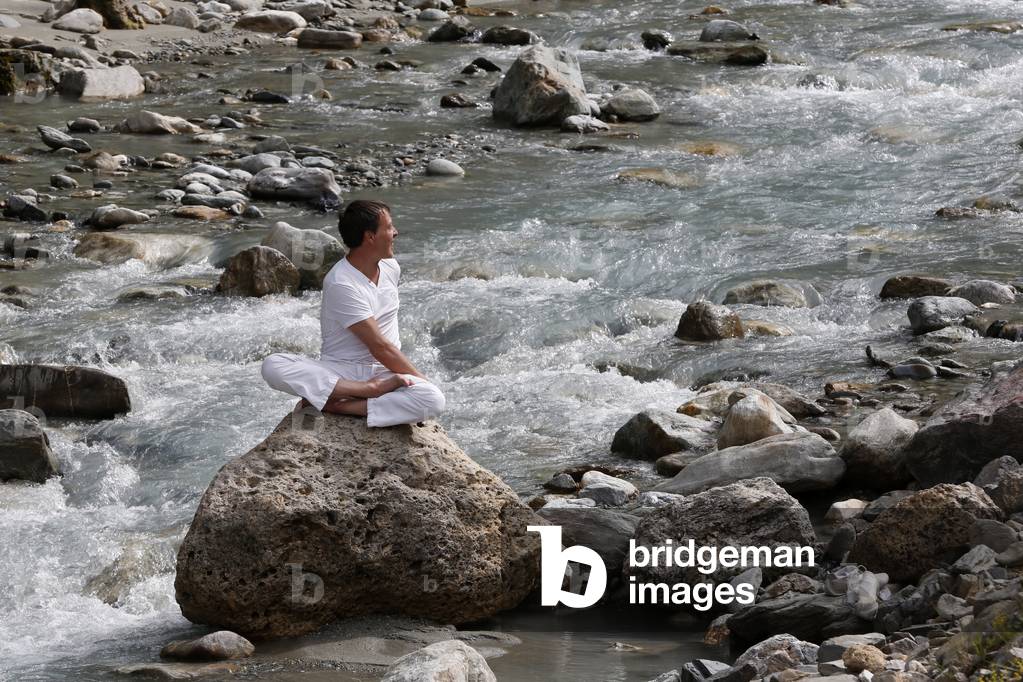 Man meditating in the lotus position, sitting on a rock by the river, Les Contamines, France