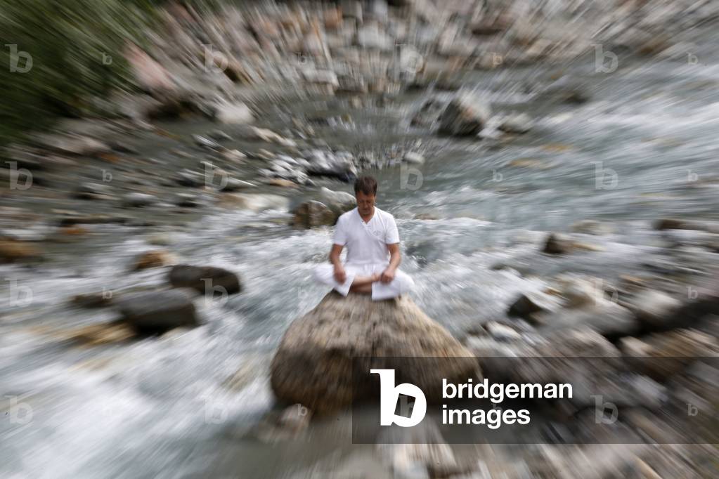 Man meditating in the lotus position, sitting on a rock by the river, Les Contamines, France
