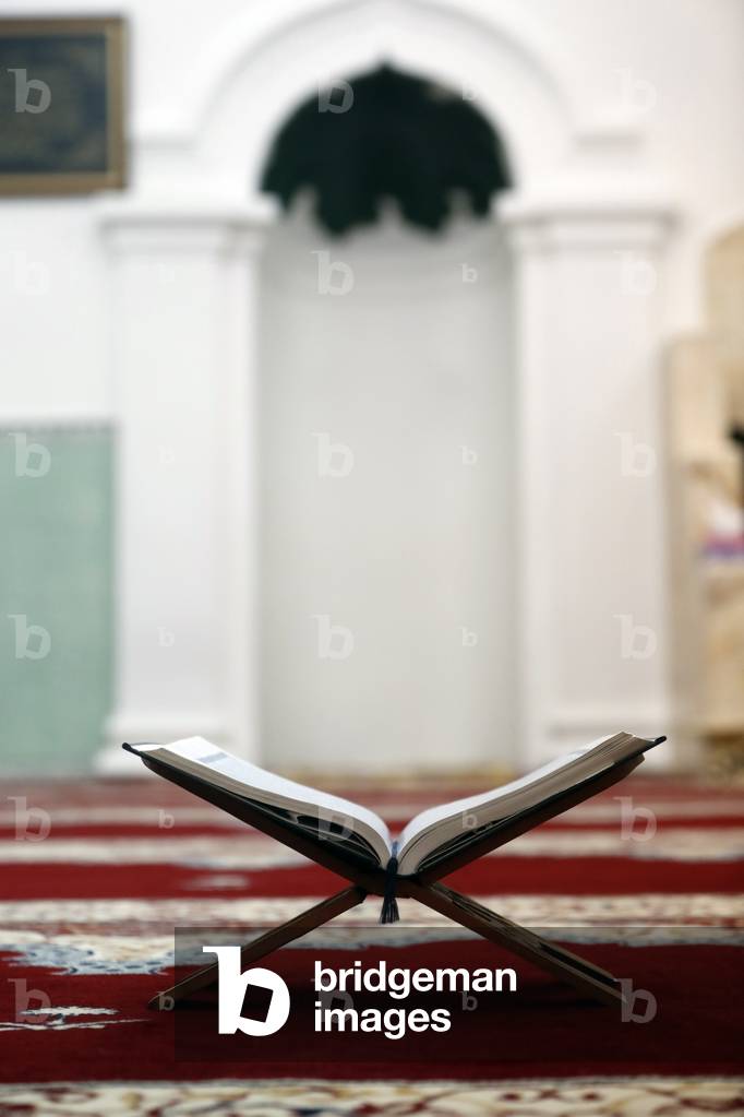 An open Holy Quran on wood stand with mihrab in background (photo)