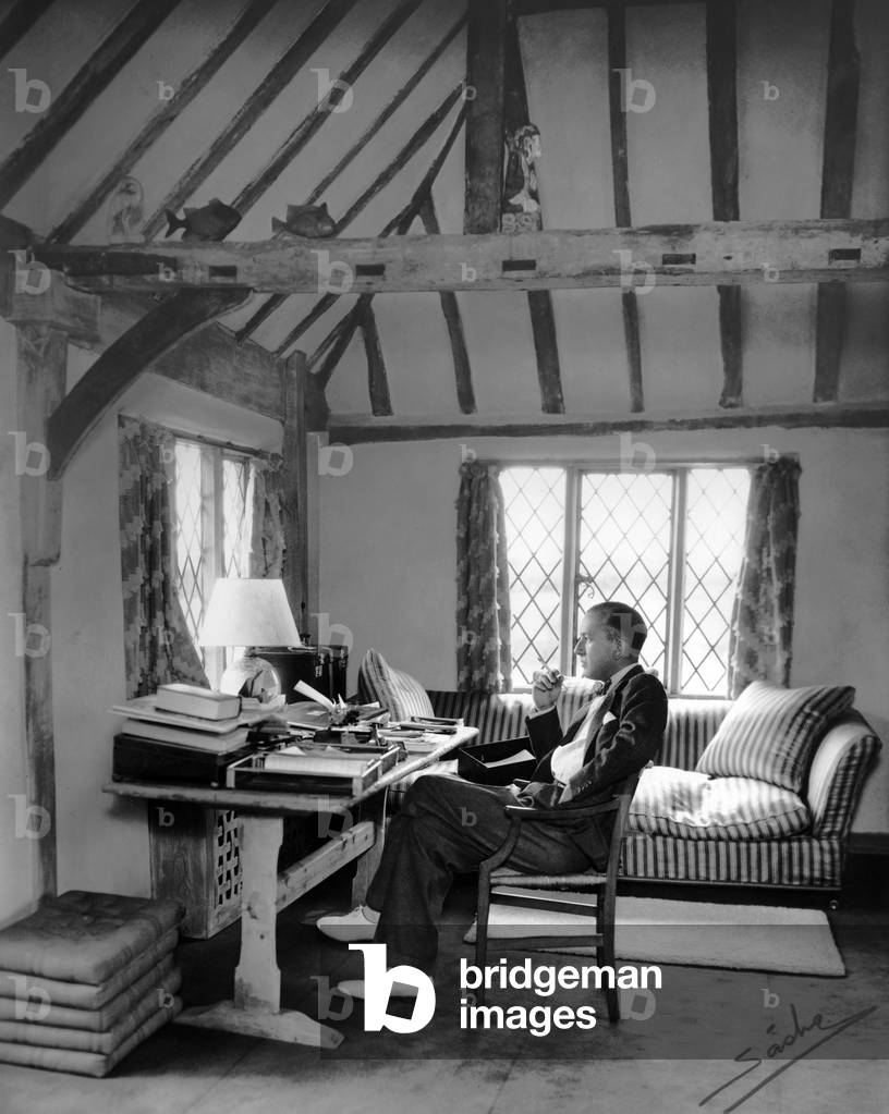 Portrait of Noel Coward seated at a desk, smoking