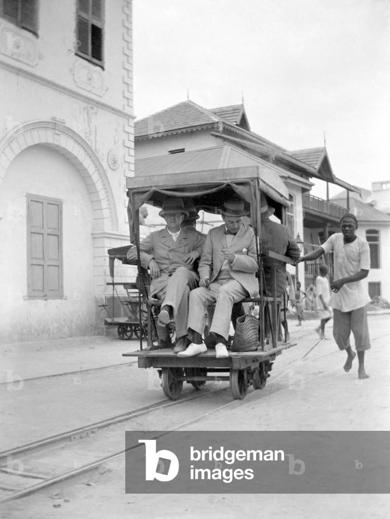 African pushing Europeans on a cart laying on small rails. photography around 1920