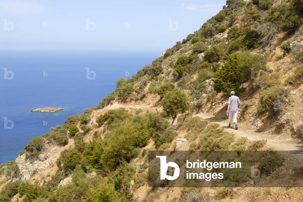 Woman hiking along a steep coastal cliff trail; looking over an island and the blue sea; descending from Moutti tis Sotiras mountain; Baths of Aphrodite; Akamas; Southern Cyprus; Republic of Cyprus; Mediterranean Sea; Europe