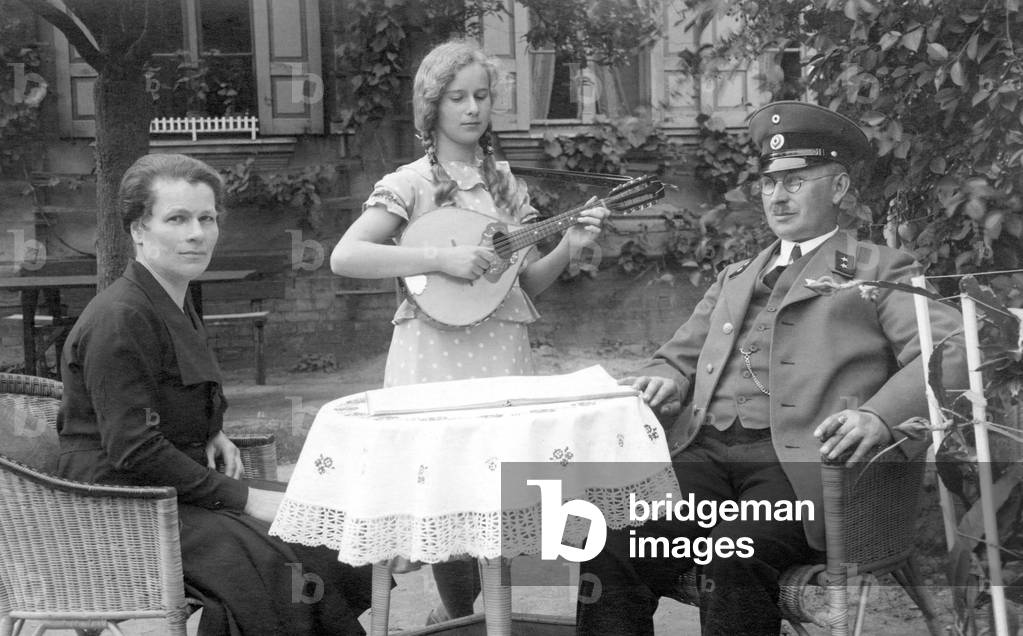 Family portrait of the German bourgeoisie gathered around a table, the little girl plays mandolin for her parents, has austere attitude, the father probably engages in the First War. 1915 approximately Historic photo, parents with daughter who is playing music, ca. 1915