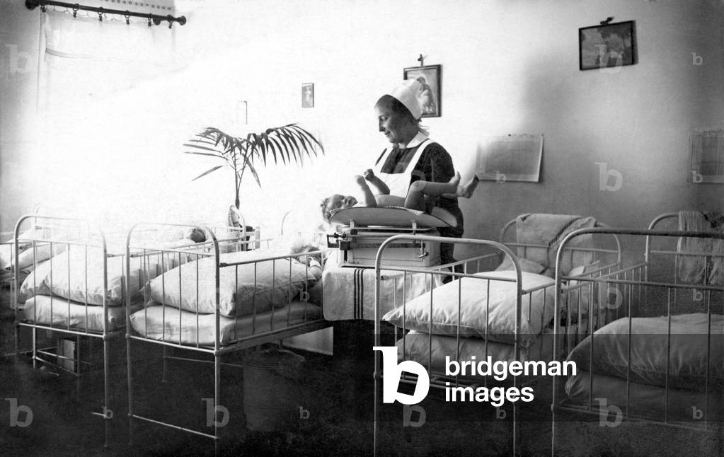Nurse and baby in a hospital. The woman weighs the newborn on a scale. Photography 1910