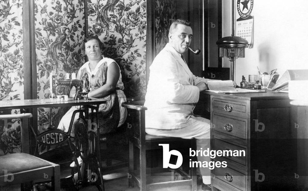Couple at home: the woman in apron in front of her sewing machine and the man in her office smokes a pipe. Photography around 1920.