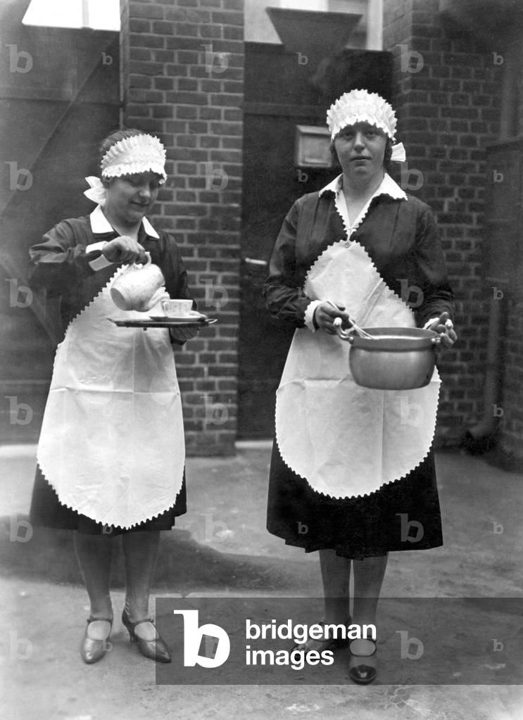Two domestic women have the kitchen: one prepares the tea and the other makes a cake. Photography around 1920