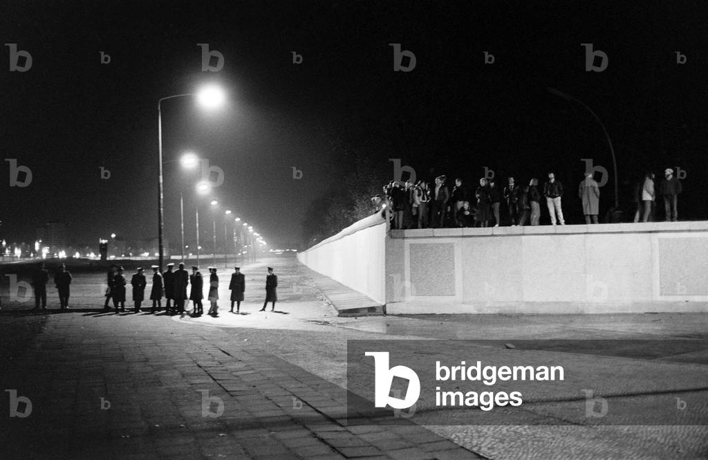 GDR border guards standing in the no man's land at night while West Berliners are standing on the wall, 9 November, 1989 (b/w photo)