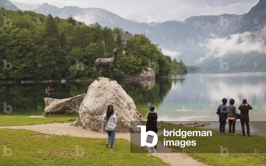 Lake Bohinj (Bohinjsko jezero), Triglav National Park, Upper Carniola, Slovenia. Visitors admiring the lake in the early morning at the site of the statue of Zlatorog.  Zlatorog is a mythical chamois with golden horns immortalized by German poet Rudolf Baumbach and the writings of Slovenian politician and natural scientist Karel Deman, also known as Dragotin Deman and Karl Deschmann.