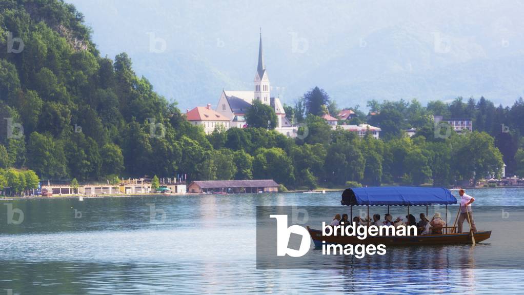 Bled, Upper Carniola, Slovenia.  Church of St Martin seen across Lake Bled.  Tourists enjoying excursion on boat.