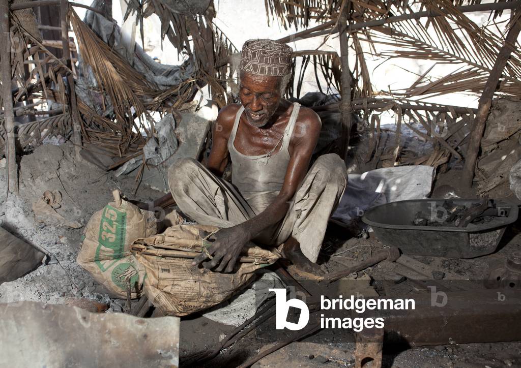 Blacksmith sitting on the floor with kofia sweating while he forges nail, Lamu, Kenya, Africa (photo)