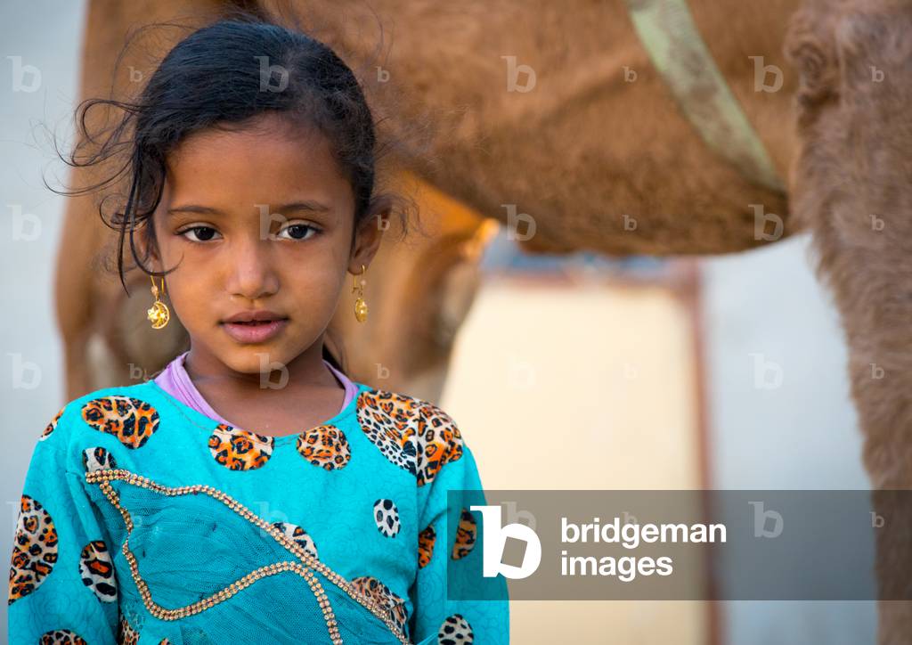 Young girl in blue dress in front of a camel, Qeshm Island, Salakh, Iran (photo)