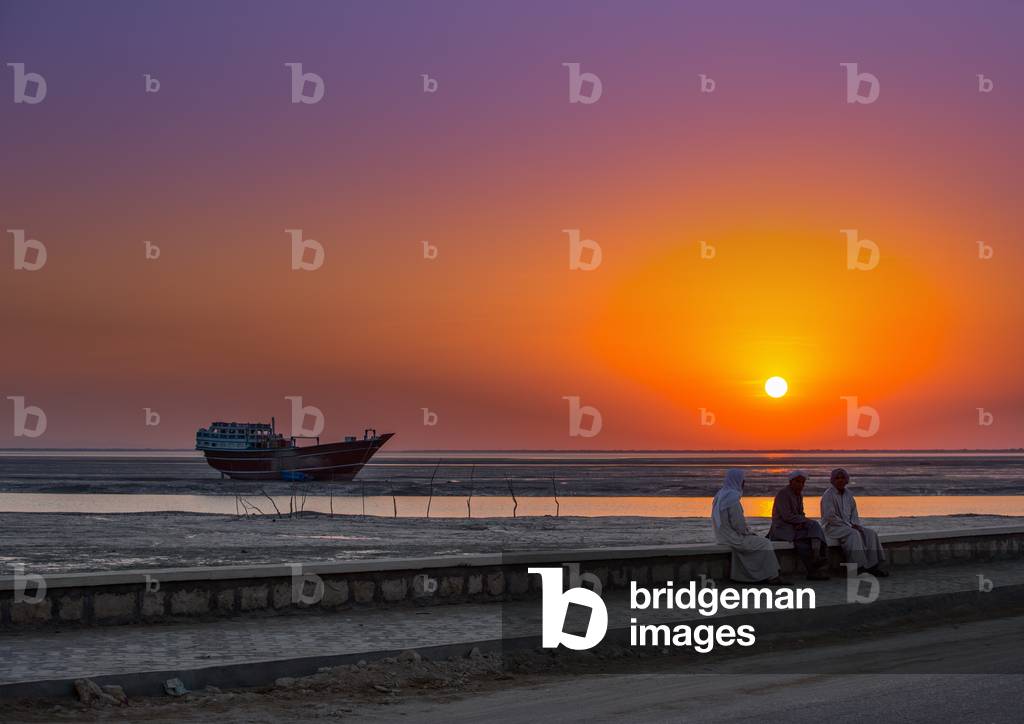 Men Dressed In Traditional Dress, Sitting On A Bench In Front Of A Sunset And A Dhow Boat, Qeshm Island, Laft, Iran, 2015 (photo)