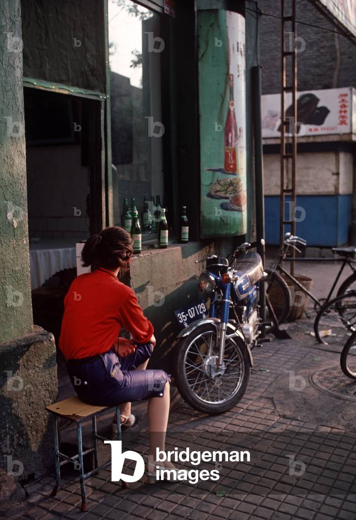 Chinese woman sitting infront of restaurant bar with food and drink painted advertising sign in Harbin, Heilongjiang Province, China (photo)