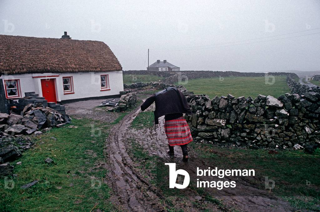 Aran Islander Wearing Traditional Red Skirt Making Her Way To Thathched Cottage During Heavy Atlantic Rain On Inishmore, Aran Islands Off The Coast Of Galway, Ireland. Referred To By W. B. Yeats When He Suggested That Playwright John Millington Synge Visit Visit The Aran Islands.  (photo)