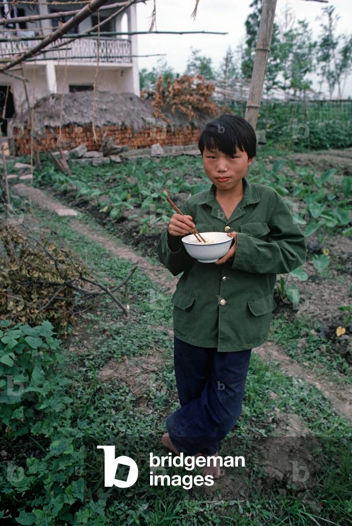 Child eating lunch of noodles with chopsticks on commune vegetable plot, Zhejiang Province, China (photo)