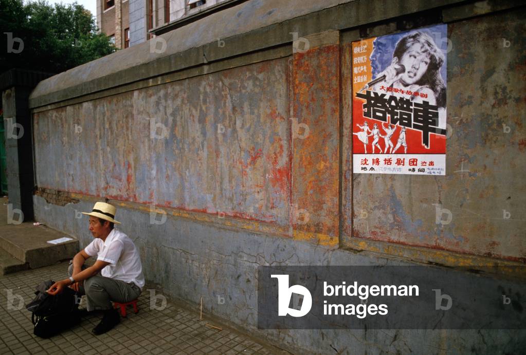 man crouching down next to wall with pop music poster, Shenyang, China. (photo)